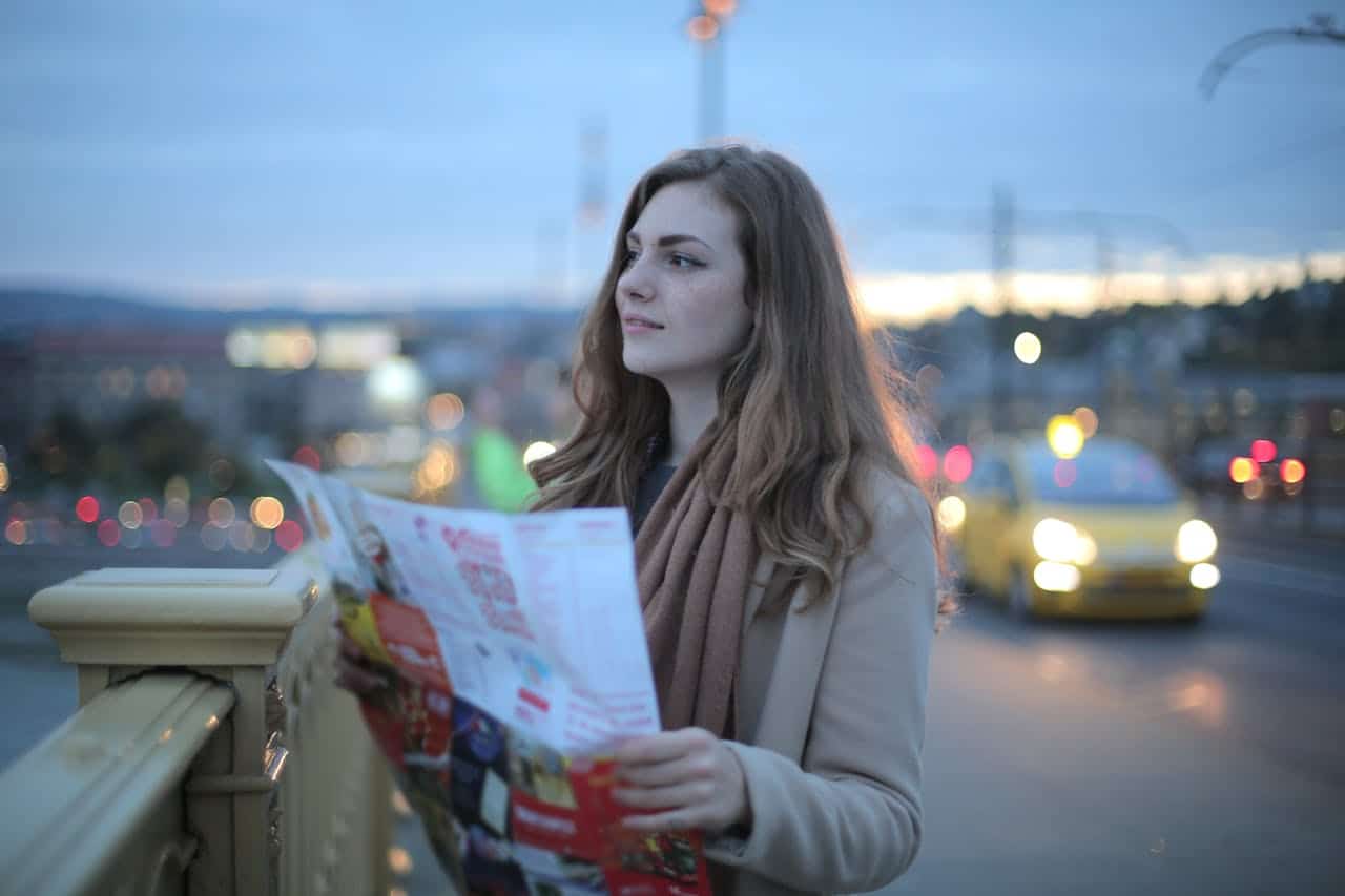 Mujer viajera en ropa de abrigo de pie en la calle con un mapa y mirando a lo lejos en el crepúsculo