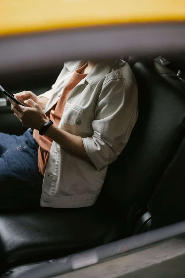 Desde arriba hombre sin rostro con chaqueta blanca y pulsera fitness sentado en el asiento trasero de un taxi y mirando el móvil