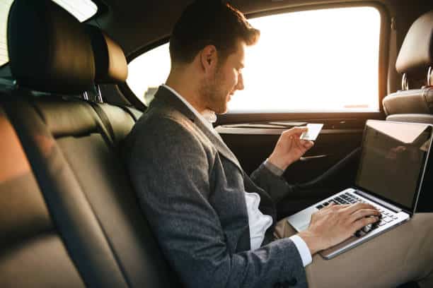 Atractivo hombre de negocios usando una tarjeta de crédito de plástico mientras está sentado con un ordenador portátil en la parte trasera de un coche.